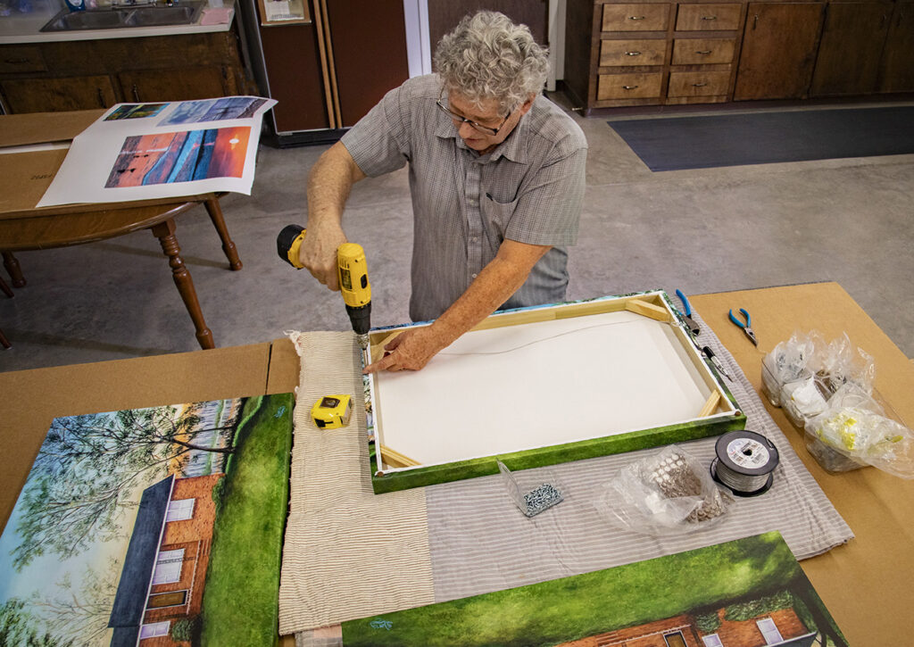 Man assembling a framed picture using a drill on a table with art supplies and other artworks in a workshop setting.