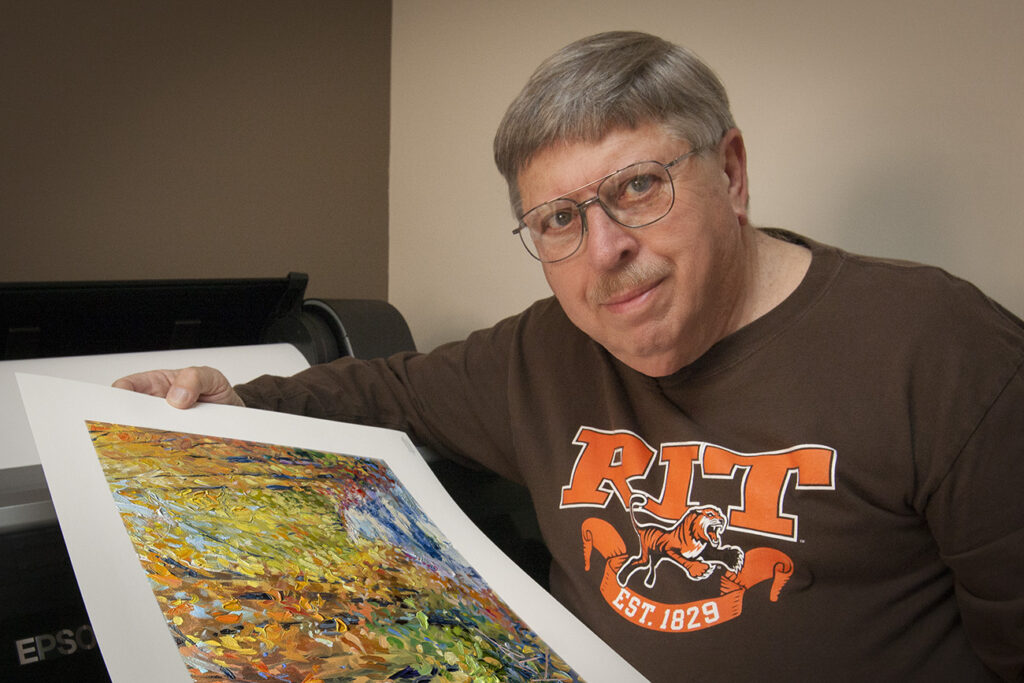 A man in glasses holds a colorful painting, wearing an RIT shirt, standing next to a printer in a room with neutral walls.